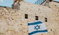 Eine israelische Flagge weht vor einer alten Steinmauer mit Fenstern und einem Turm im Hintergrund. Der Himmel ist teilweise bewölkt. (Text KI generiert)